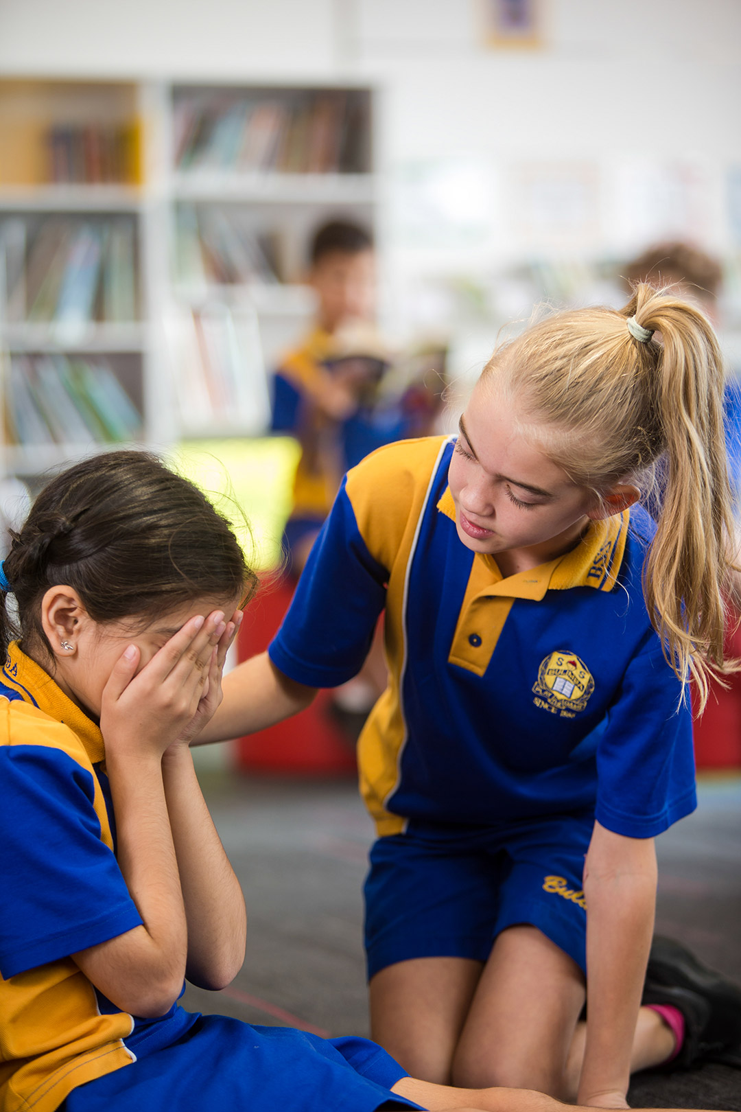 Student seated with alone on bench with her back to the camera while other students play on the playground 