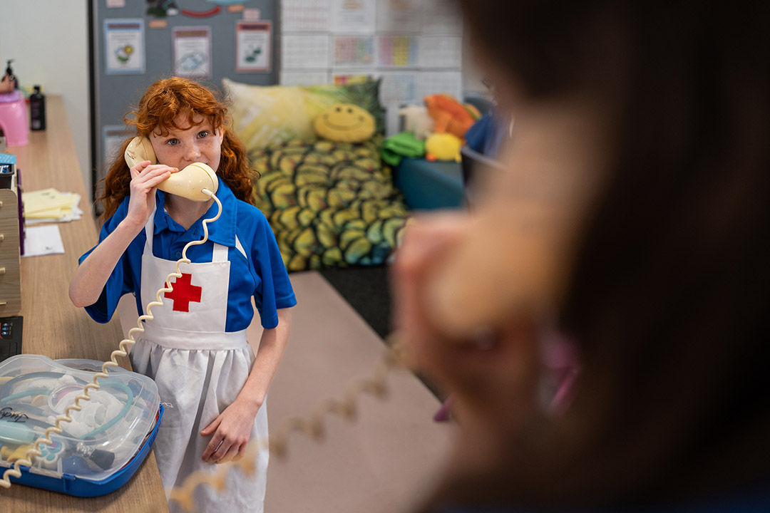 A student in a nurse apron in a classroom, holding a toy telephone to her ear.