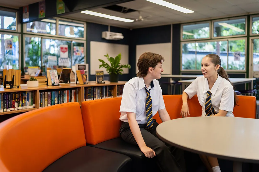 Two school students in uniform sitting together and talking in a school library