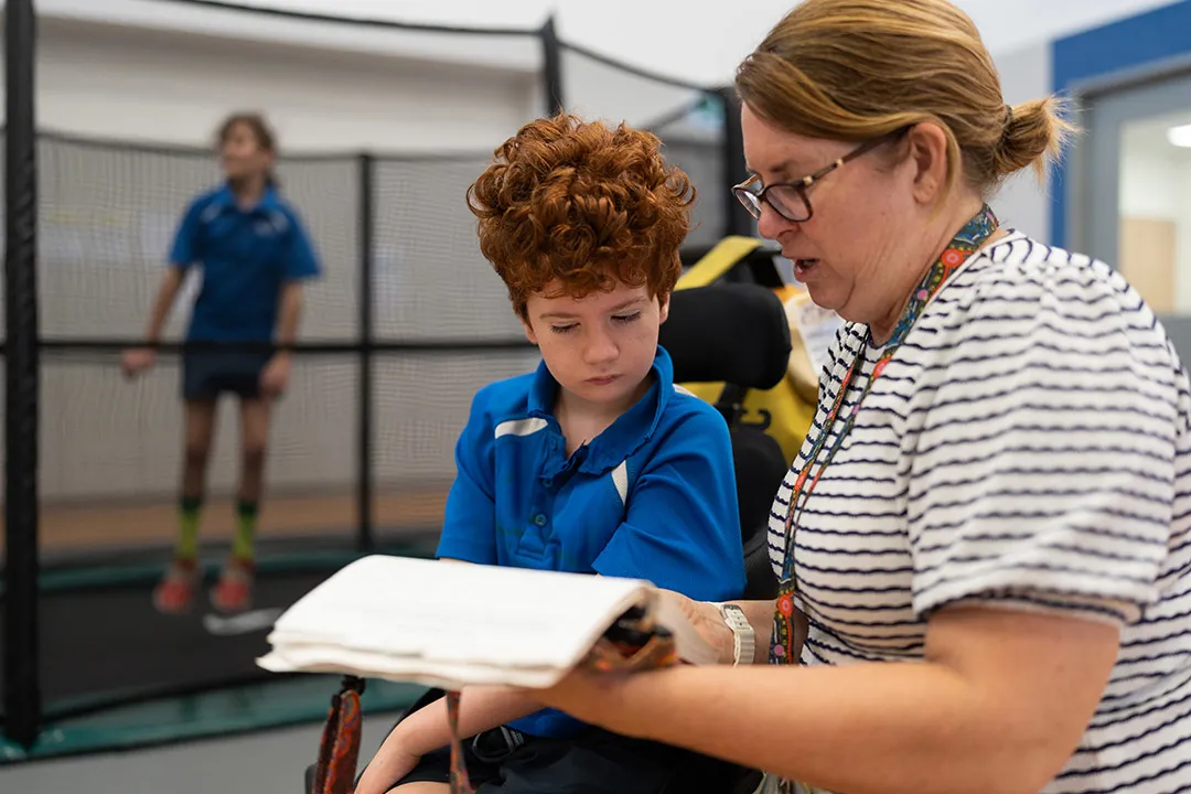 A primary school child in a wheelchair is looking at a booklet with his teacher guiding him. There is another child in the background jumping on a trampoline.
