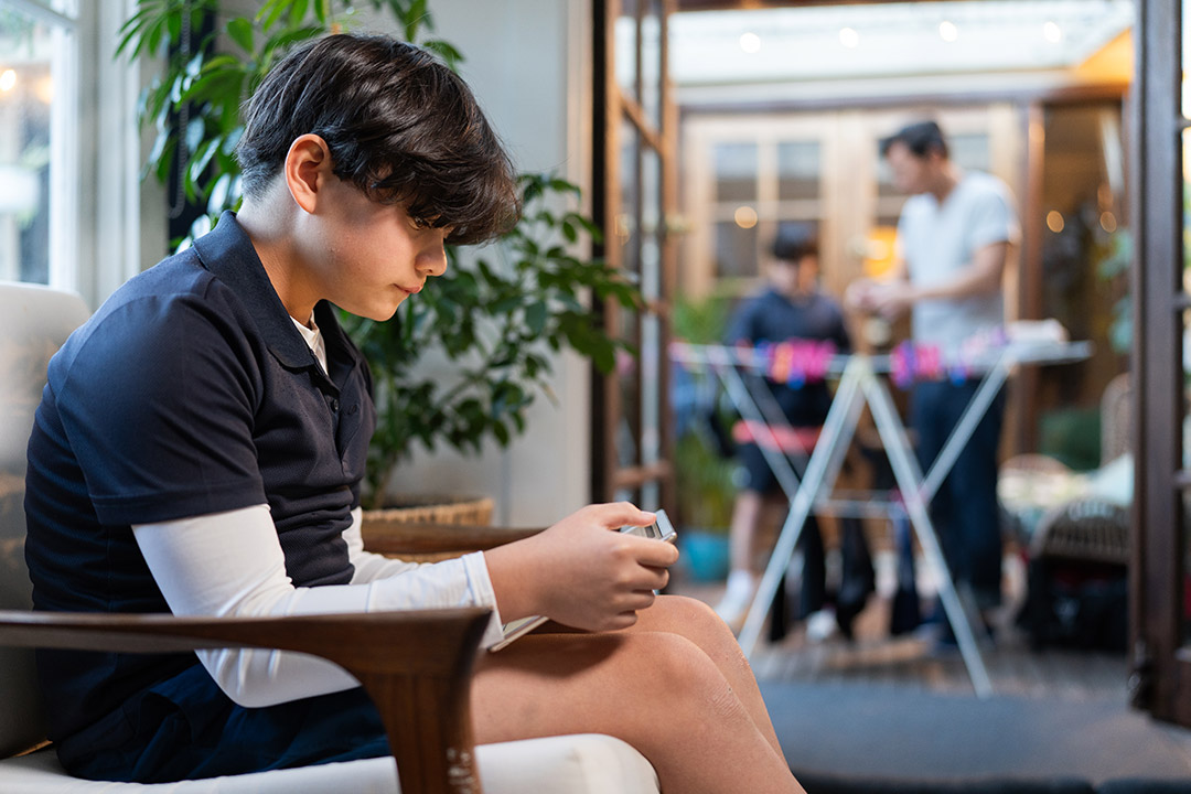 Student sitting on chair in house playing on device. Parent and younger sibling handing washing out in background.