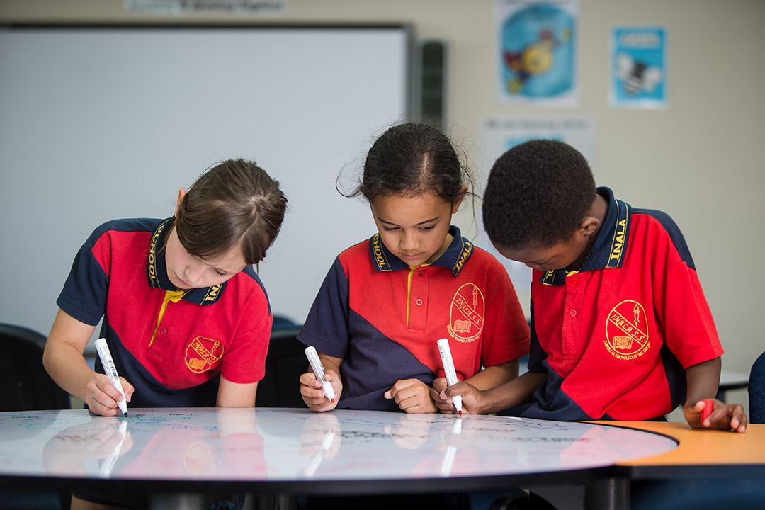 Three primary students wearing school uniform writing on a whiteboard. 