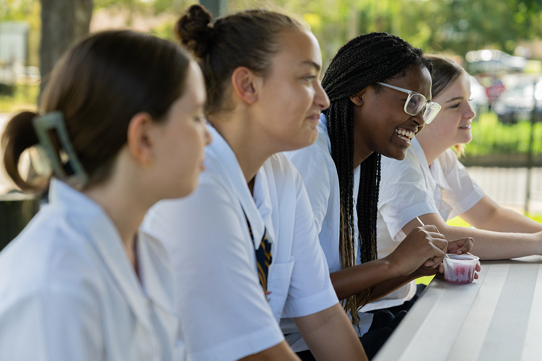A group of four secondary school students in uniform sat at a bench together talking and laughing.