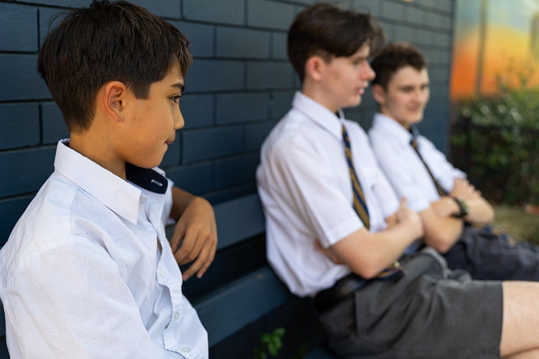 Three boys in school uniform sitting on a bench. One is sitting further away and looks withdrawn.