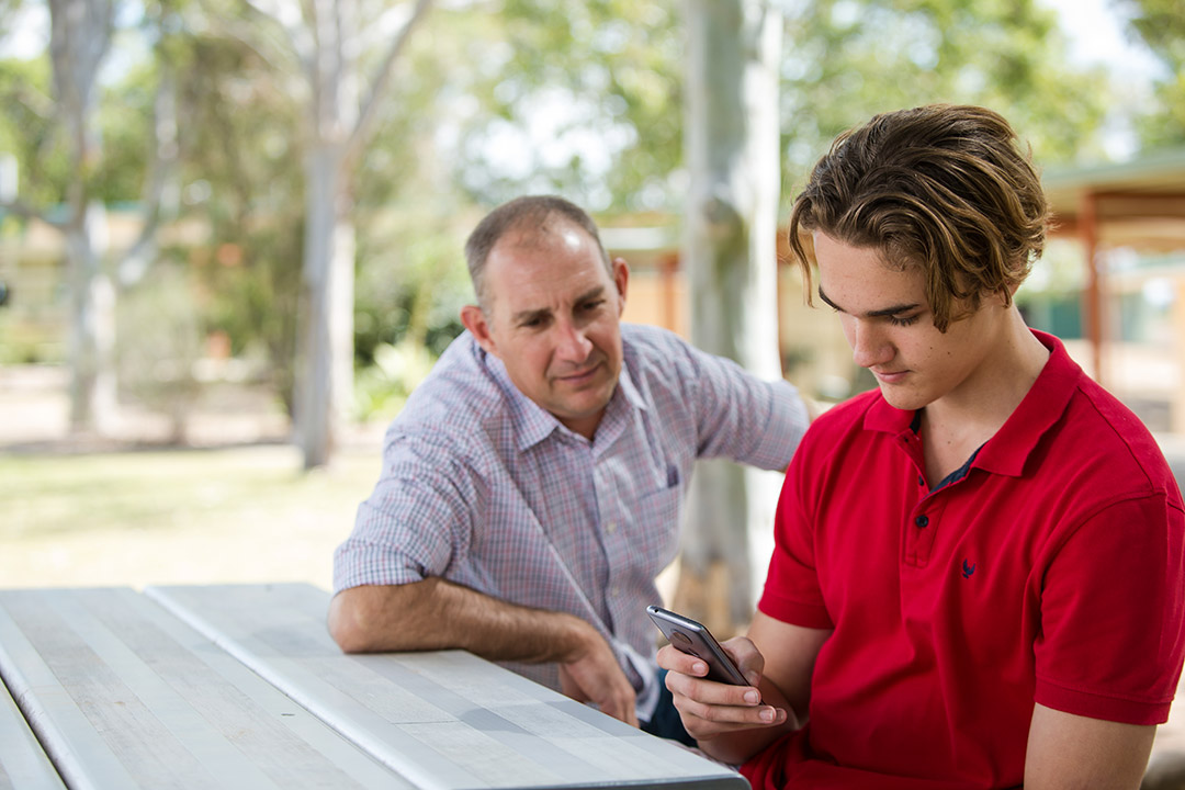 A teenager on his phone seated on a bench outside next to an older man