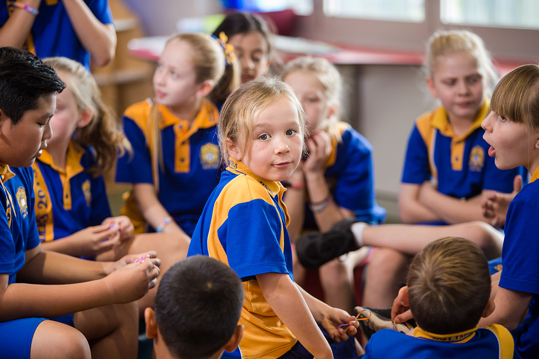A group of primary school students sitting together in conversation. One of them, looks isolated towards the camera.