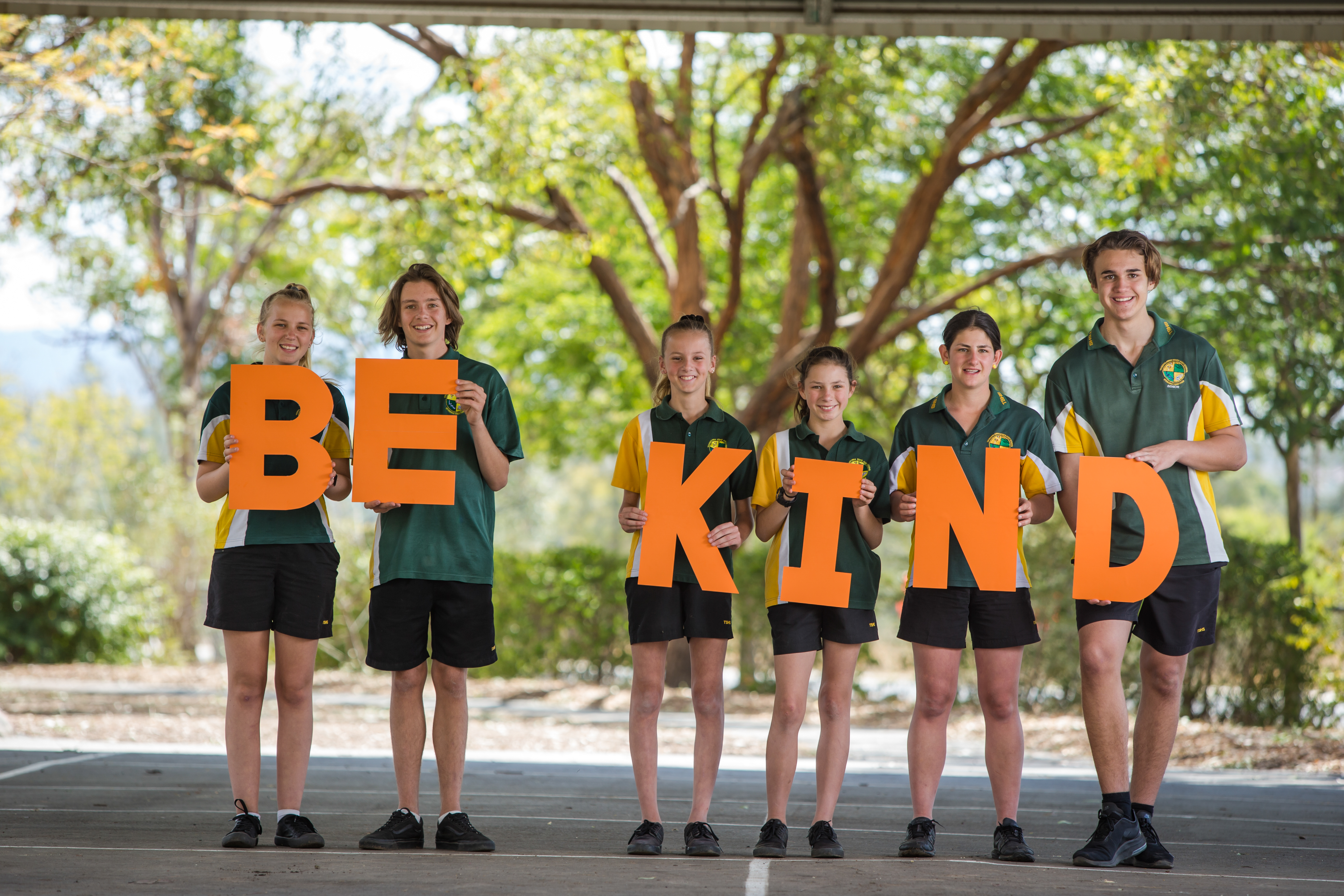 Six school students in uniform, each holding a letter to spell out 'Be Kind'